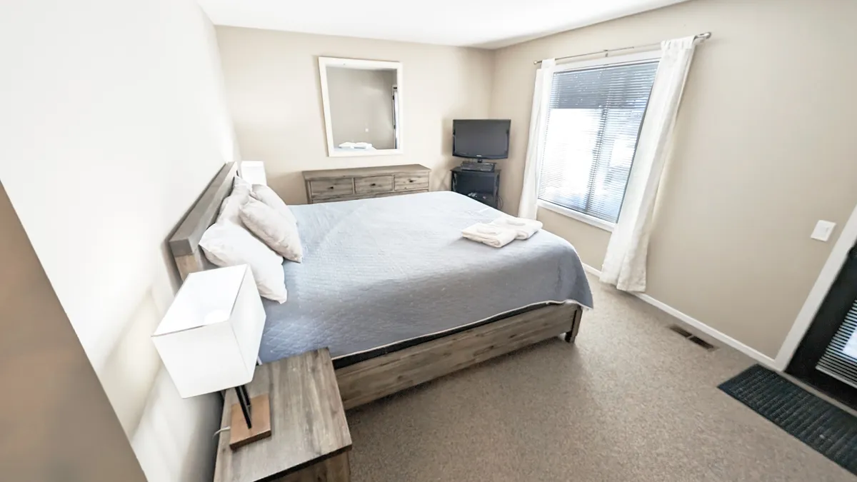 A serene bedroom with a king-sized bed, a dresser, mirror, window letting in natural light, and a flat-screen TV on the wall. The room exudes comfort and calm, suggesting a restful retreat ideal for recharging.