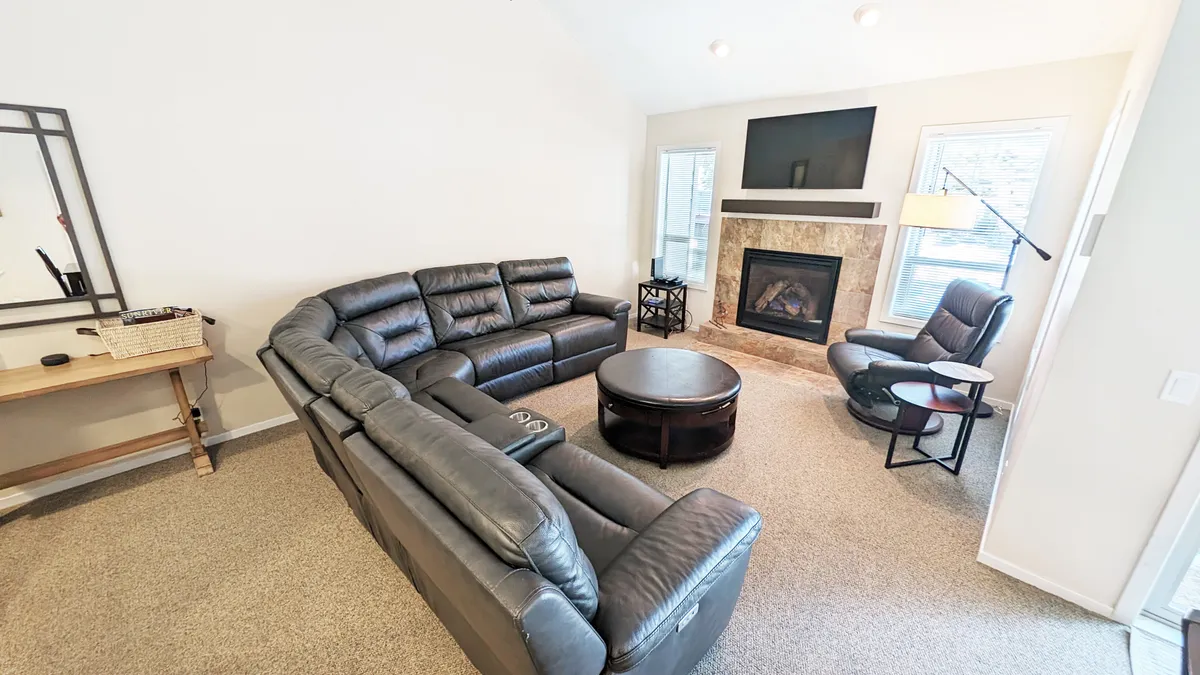 The image showcases a bright, welcoming living room with a plush sectional sofa, a cozy fireplace, a stylish coffee table, and an inviting armchair. The space is filled with natural light and designed for gathering, with ample room for relaxation, movie nights, and enjoying time together.
