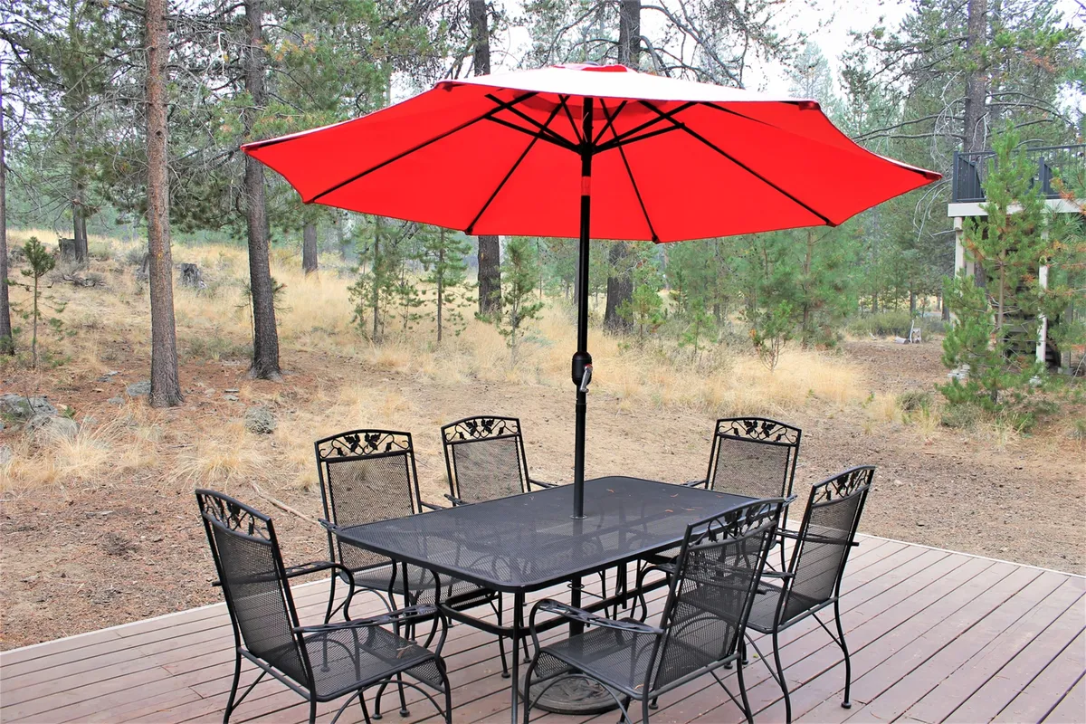 An inviting patio scene with a red umbrella shading a black dining set, all framed by lush trees. The setting is clearly designed for outdoor meals and gatherings, creating a cozy transition between indoor comfort and the beauty of nature.