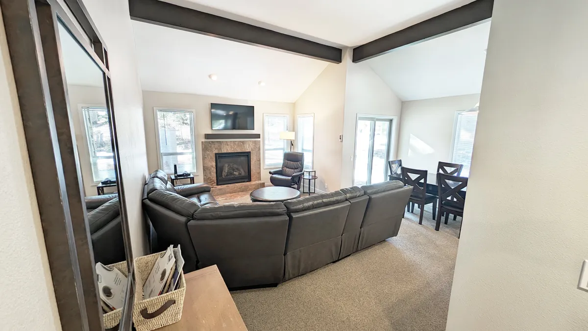 A spacious living and dining area with a leather sectional sofa, fireplace, dining table, and large windows letting in abundant natural light. The windows frame the natural surroundings, reinforcing the connection to Sunriver's outdoor beauty.