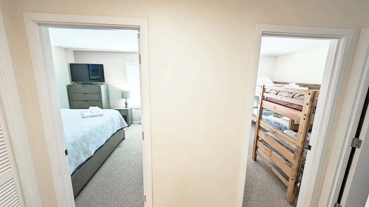 A family-friendly bedroom featuring a standard bed with white linens, a bunk bed with cozy bedding, a TV, and a nightstand with a lamp. The space is playful yet comfortable, offering versatility for families or groups.