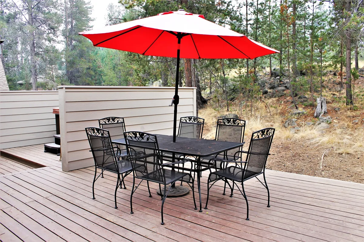A relaxed exterior view featuring a red umbrella over a black metal table, set on a wooden deck amidst tall trees. The space is ideal for family gatherings, barbecues, or morning coffee.