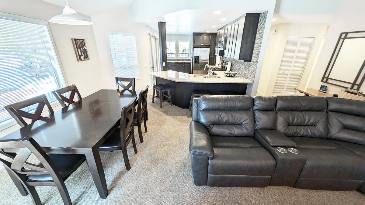 A harmonious view of the dining table, kitchen island, and sectional sofa arranged in an open layout with large windows. The design fosters a comfortable, social atmosphere for family meals, entertaining guests, and enjoying casual gatherings.