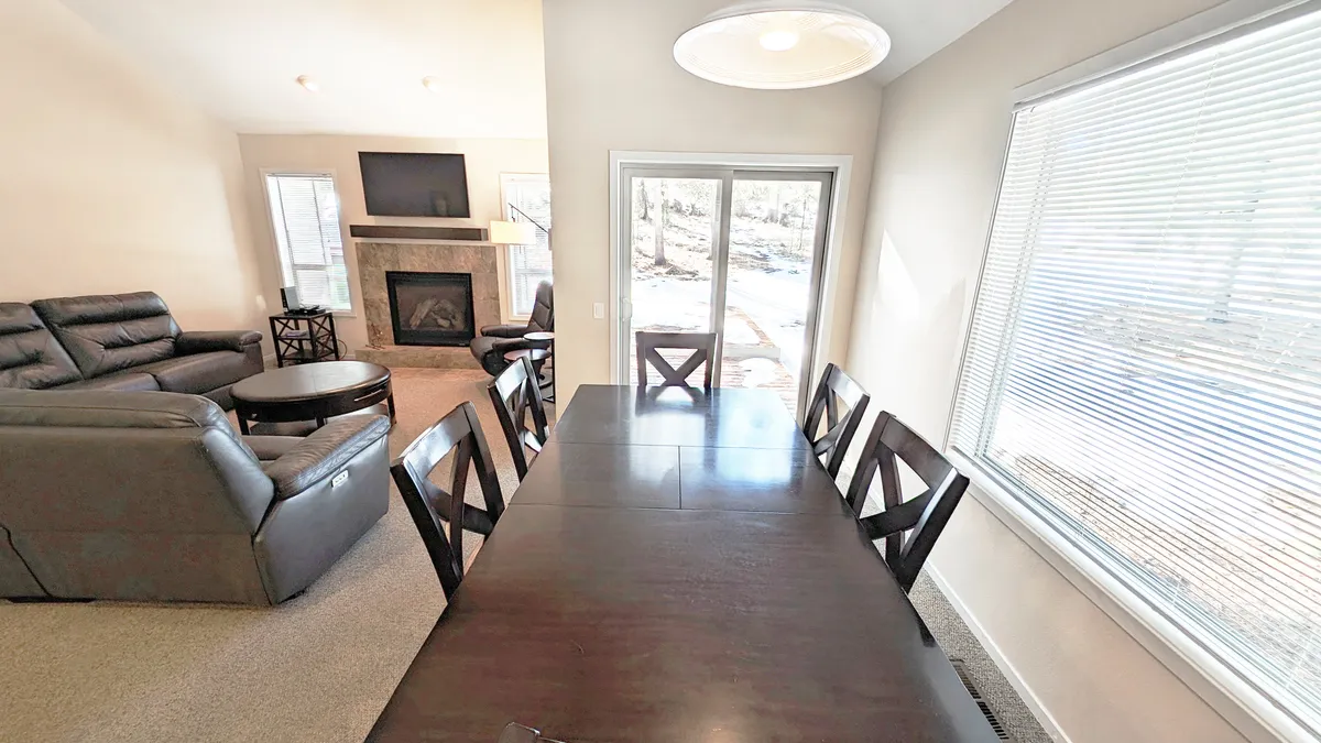 A spacious, open-plan living and dining space featuring a large dining table, comfortable seating area, fireplace, and expansive windows that fill the room with natural light. The setup is ideal for family meals, game nights, and relaxed evenings by the fire.