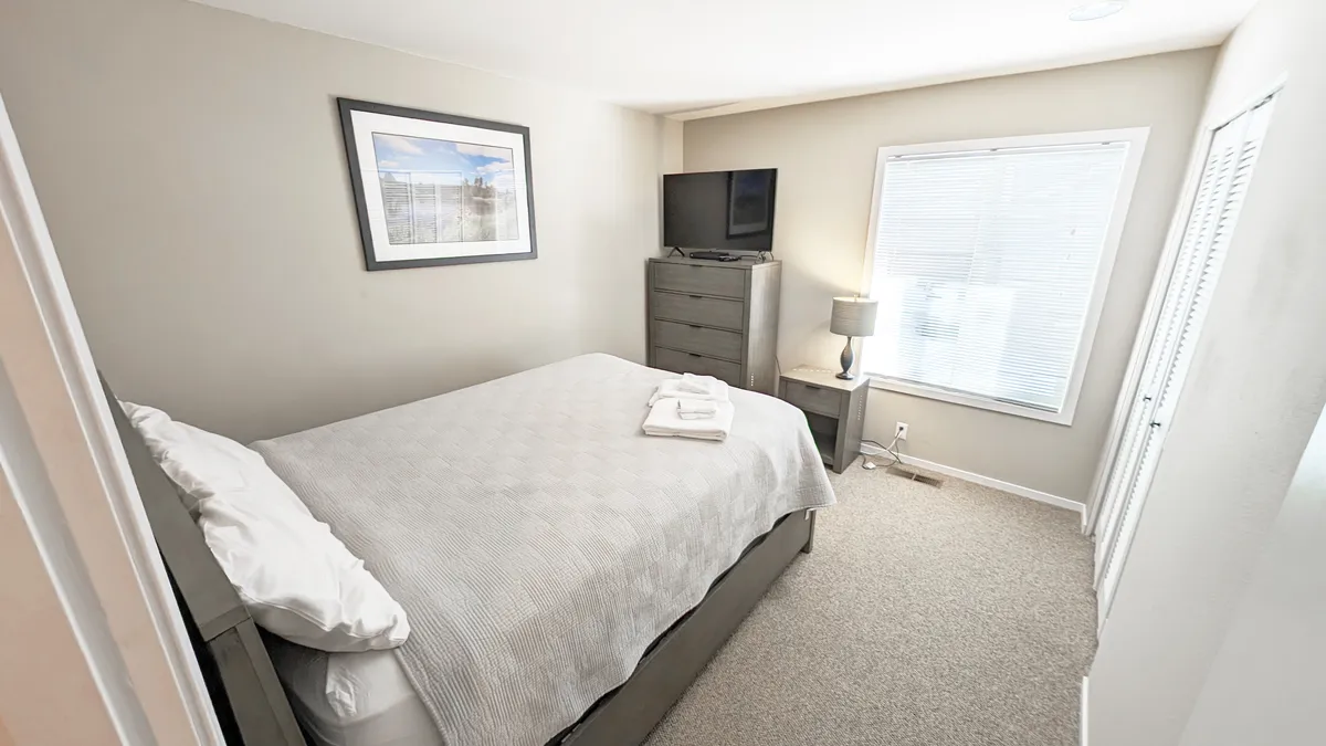 A peaceful bedroom with a large bed, window allowing in soft daylight, a TV atop a dresser, and a lamp for evening ambiance. The design is simple, quiet, and inviting—ideal for couples or grandparents.